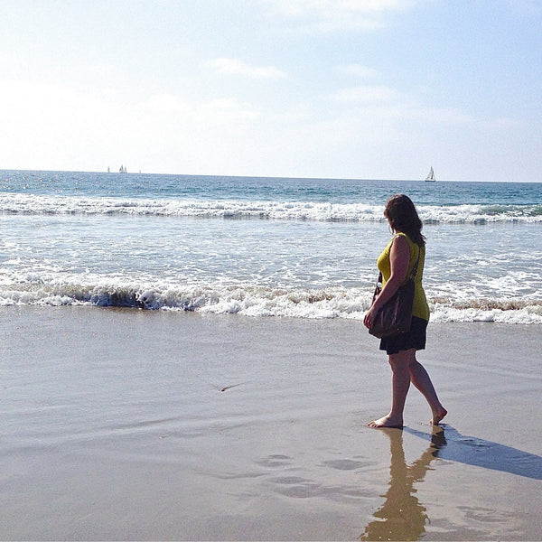 portrait of jeweler for Foamy Wader from over her left shoulder, wading in the surf with rolling waves approaching. There are sailboats on the horizon and the artist's reflection can be seen in the wet sand.