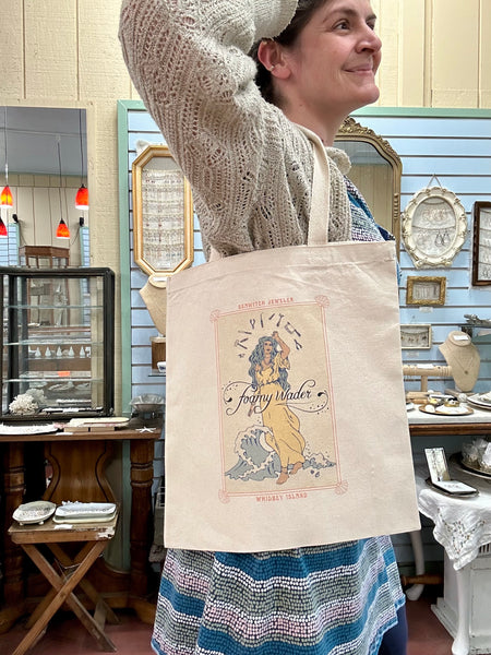 Tote bag with Foamy Wader logo, text that reads "seawitch jeweler" and "whidbey island", and art nouveau-style tarot card like imagery on a square cotton canvas bag. Tote is modeled by a woman facing screen right, wearing an ivory woven sweater with her arm raised.