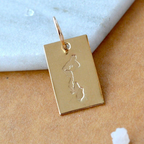 Rectangle charm with Whidbey Island impressed in gold. Displayed on a white marble surface on top of a brown textured backdrop.