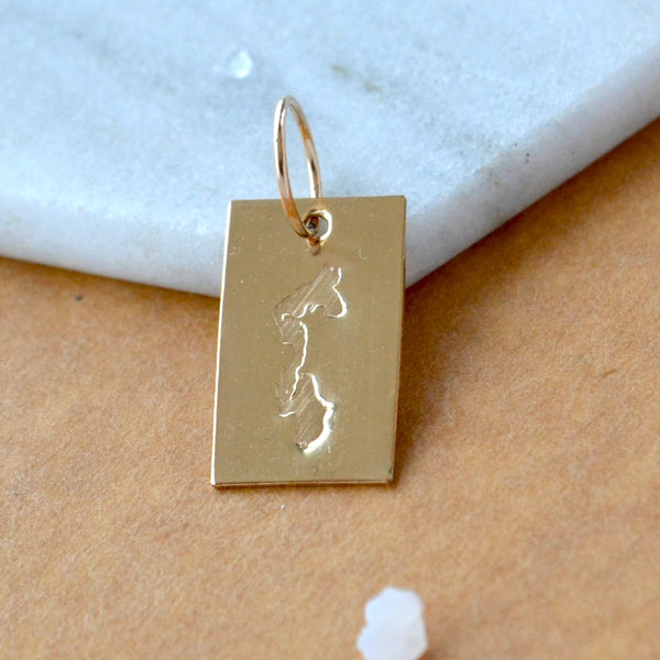 Rectangle charm with Whidbey Island impressed in gold. Displayed on a white marble surface on top of a brown textured backdrop.