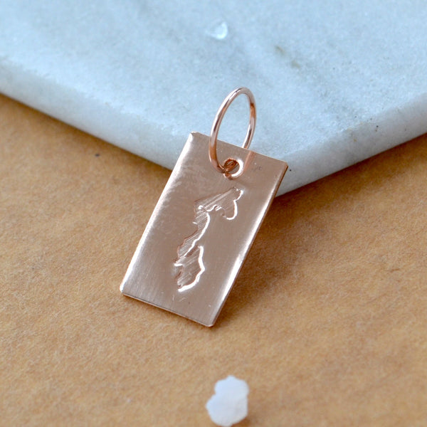 Rectangle charm with Whidbey Island impressed in rose gold. Displayed on a white marble surface on top of a brown textured backdrop.