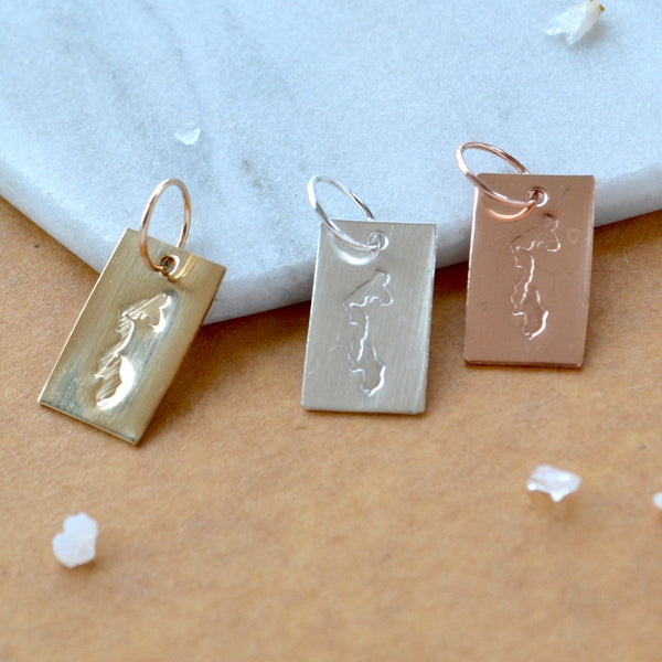 Three rectangular charms with Whidbey Island impressed in gold, silver, and rose gold. Displayed on a white marble surface on top of a brown textured backdrop.