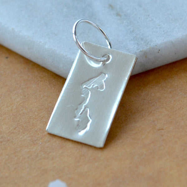 Rectangle charm with Whidbey Island impressed in silver. Displayed on a white marble surface on top of a brown textured backdrop.