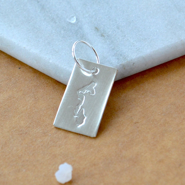 Rectangle charm with Whidbey Island impressed in silver. Displayed on a white marble surface on top of a brown textured backdrop.