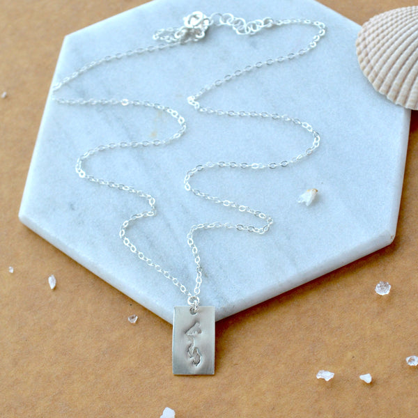 Silver necklace with a rectangular pendant of Whidbey Island on a marble surface with a shell and small stones.