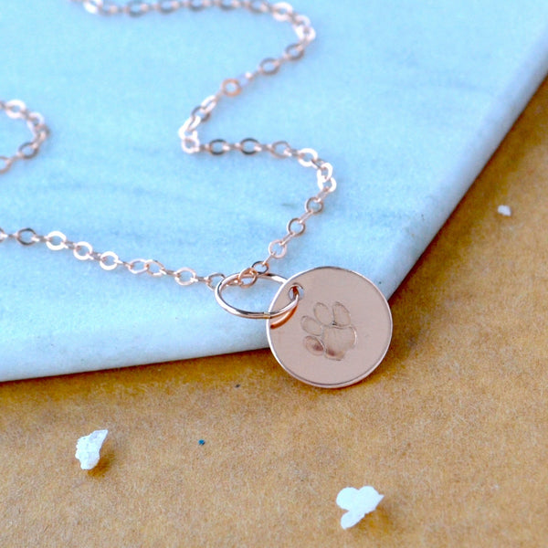 Rose gold necklace with a round dog paw charm pendant on a light blue and brown background