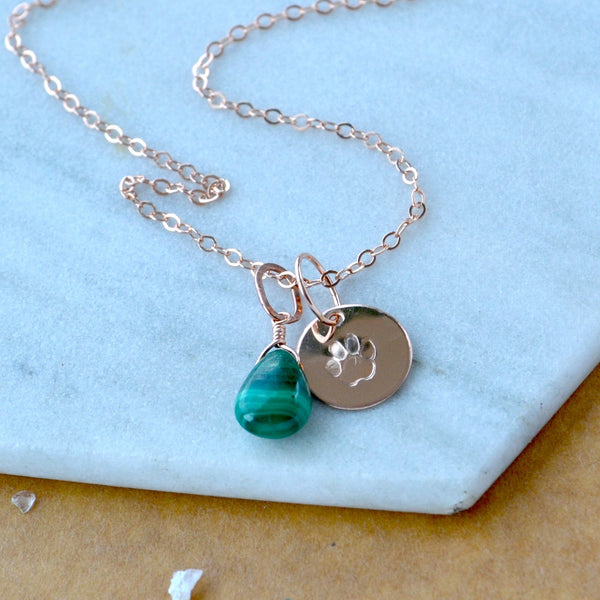 Necklace with green malachite teardrop pendant and circular dog paw charm on a light blue surface