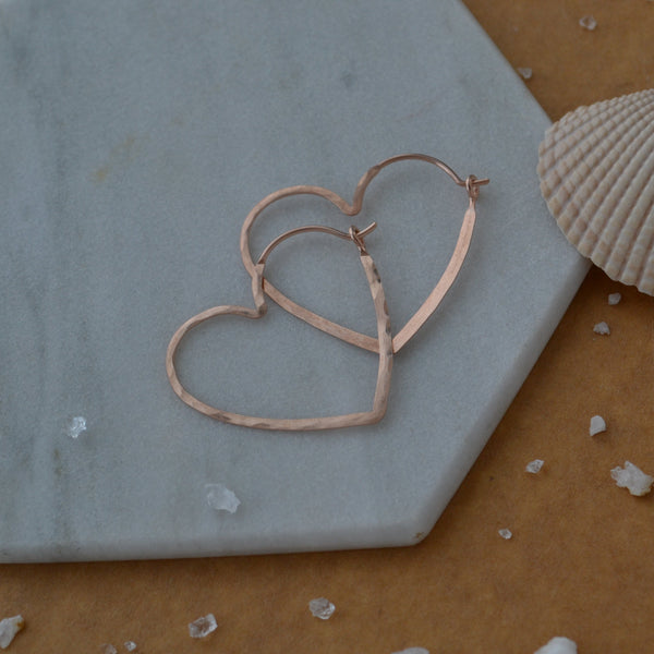 Rose gold heart-shaped earrings on a hexagonal stone surface with a shell and small stones.